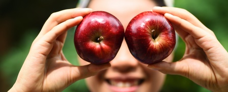 En person håller två röda äpplen framför ögonen och ler, med suddig grön bakgrund.