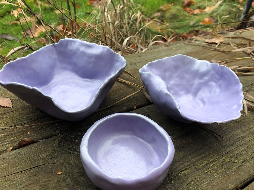 Tre handgjorda, lila keramikskålar står på ett träbord utomhus med gräs i bakgrunden.