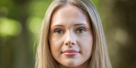Ung kvinna med långt blont hår står utomhus i grönskande miljö och tittar in i kameran.