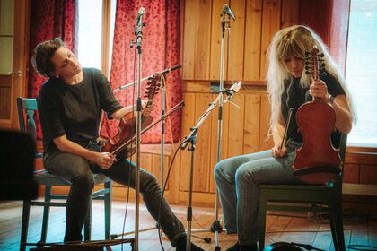 Två personer sitter på stolar och spelar nyckelharpa och fiol i en träpanelad studio med mikrofoner.