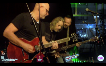 Tre män spelar elgitarr på en scen med mikrofoner och färgglada lampor i bakgrunden.