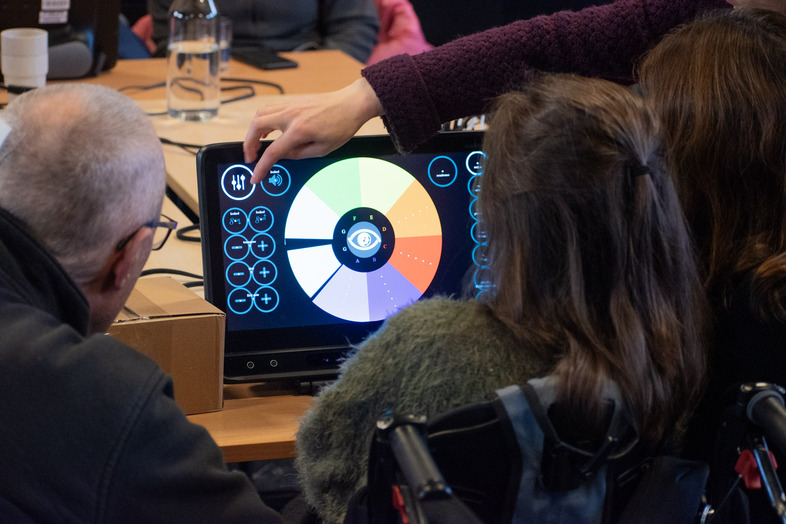 Tre personer använder en pekskärm med färgglad grafisk display vid ett bord.