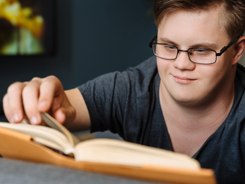 Ung man med glasögon och Downs syndrom läser en bok och vänder sida i ett vardagsrum.