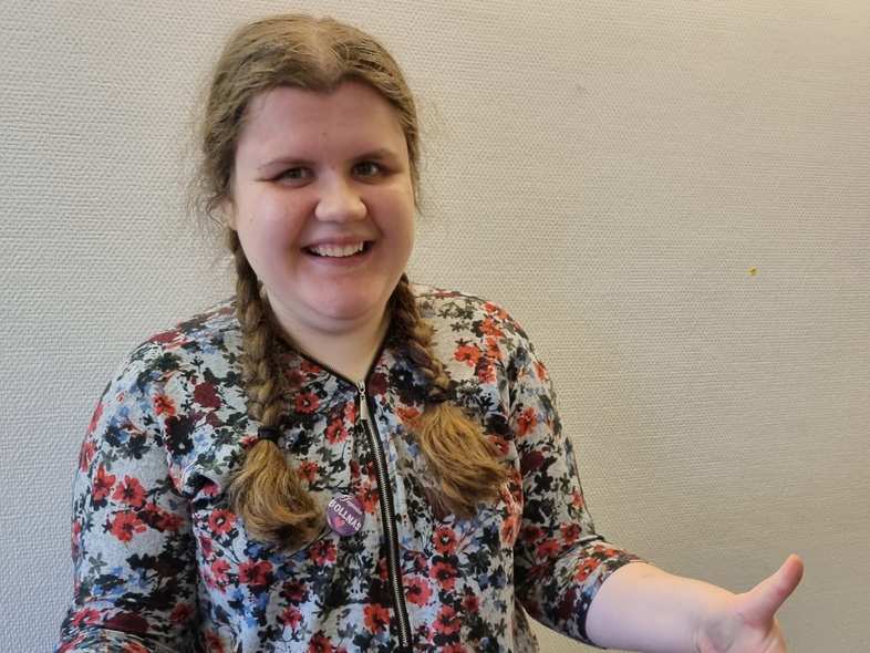 Person med flätor och blommig tröja gör tummen upp med båda händer mot en ljus vägg.