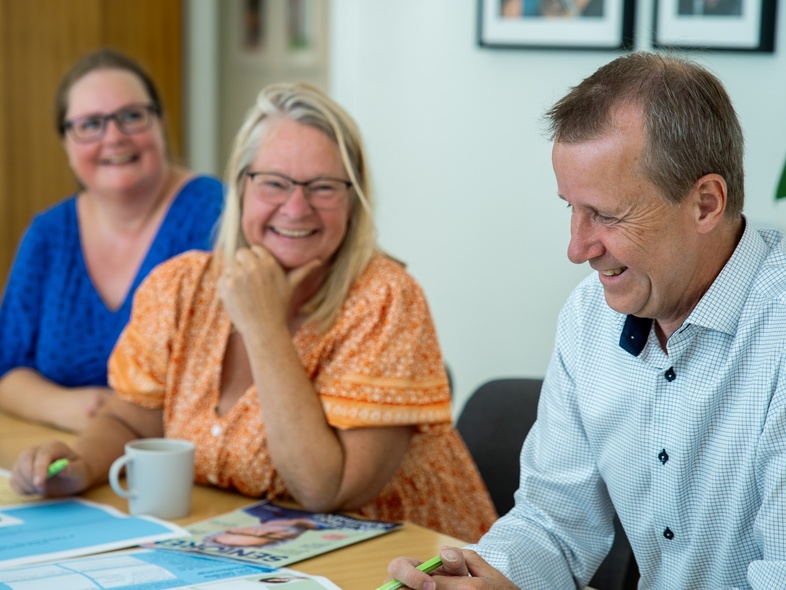 Tre personer sitter vid ett bord och skrattar under ett möte, med papper och pennor framför sig.