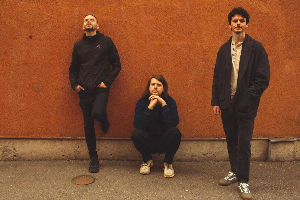 Jonatan Sirner trio poserar mot en orange vägg, en står lutad, en sitter på huk och en står rakt upp.