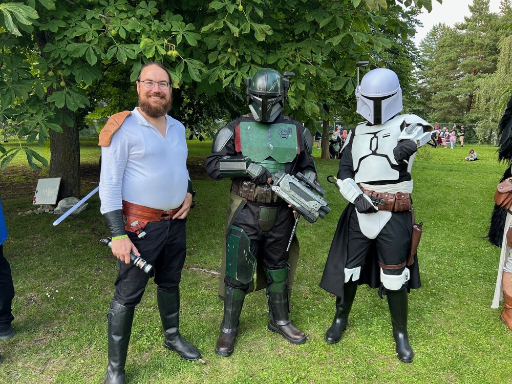 Tre personer i Star Wars-inspirerade dräkter poserar utomhus på gräs, två i Mandalorian-rustningar.