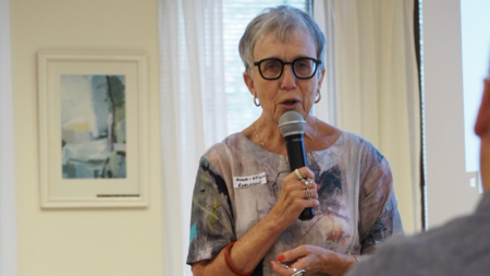 Äldre kvinna med glasögon talar i mikrofon framför en grupp inomhus.