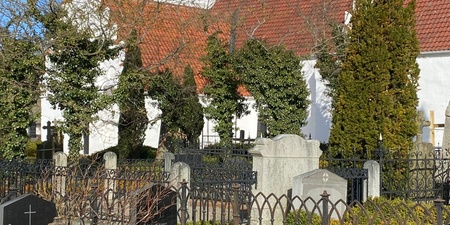 Vit kyrka med rött tak och gravstenar omgiven av järnstaket under blå himmel.