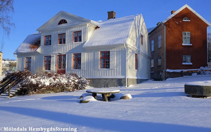 Två hus i vintrigt landskap med snö på marken och taken, ett vitt trähus och ett rött tegelhus.