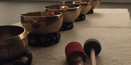 Fem tibetanska klangskålar står på rad på en matta med två klubbor framför dem i ett dämpat rum.