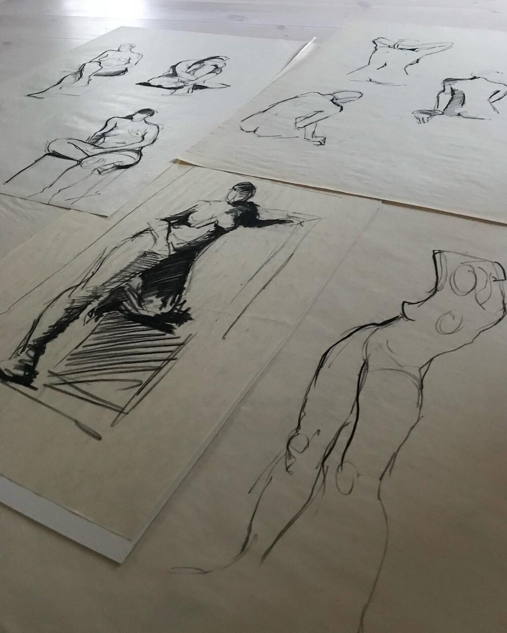 Flera skisser av nakna människokroppar i olika poser, ritade med svart bläck på papper, ligger utspridda på golvet.