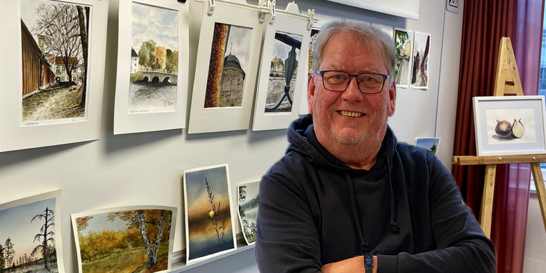 En leende man står framför en vägg med flera akvarellmålningar och konstverk.