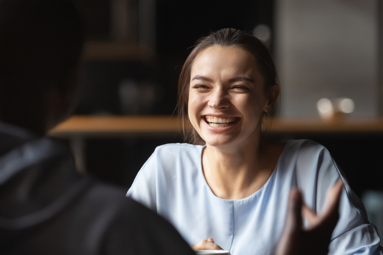 En ung kvinna med ljust hår skrattar glatt vid ett bord inomhus, mot en suddig bakgrund.