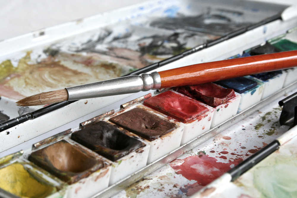 En närbild av en akvarellfärglåda med en pensel vilande ovanpå färgerna.