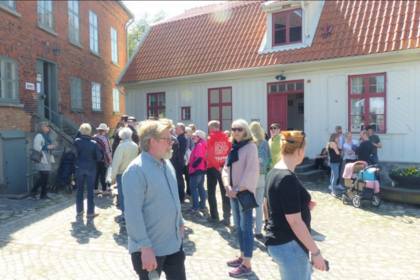 En grupp människor står på en kullerstensgata framför äldre hus i solsken.