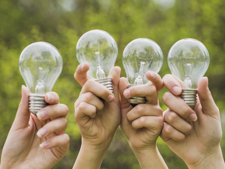 Fyra händer håller upp varsin glödlampa utomhus med grönskande bakgrund.