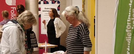 Två kvinnor står vid ett informationsbord med broschyrer på en mässa eller utställning inomhus.