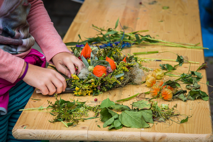 Ett barn arrangerar blommor och blad på ett träbord, omgiven av färgglada växter och löv.