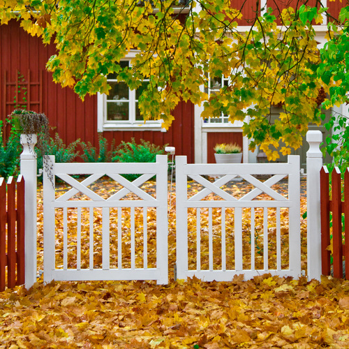 Vit trägrind framför ett rött hus, omgiven av höstlöv och träd med gula blad.