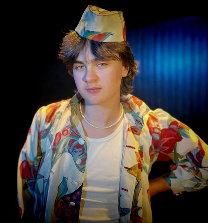 Ung person med färgglad skjorta och hatt poserar mot mörk bakgrund.