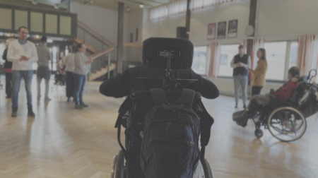 Person i rullstol i förgrunden, flera personer och en annan rullstolsburen i bakgrunden i en ljus sal.