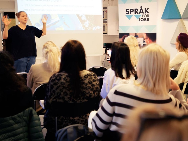 En kvinna håller en presentation inför en grupp sittande personer i ett klassrum med skylten 'Språk för jobb' synlig.