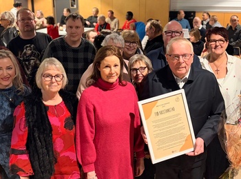 En grupp människor står samlade inomhus, en person håller ett diplom och en annan en blomsterbukett.