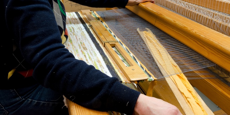 En person väver tyg på en stor vävstol i ett ljust rum. Händerna arbetar med trådarna.