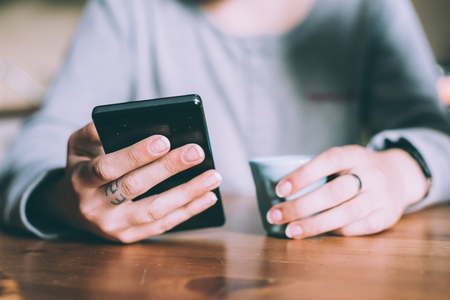 En person håller en smartphone i ena handen och en kopp i den andra, sittande vid ett träbord.