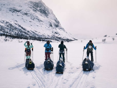 Fyra personer med pulkor och stavar står på ett snötäckt fält framför ett berg i vinterlandskap.