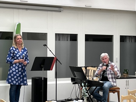 En kvinna står vid en mikrofon och en man sitter vid ett keyboard, båda inomhus i en ljus lokal.