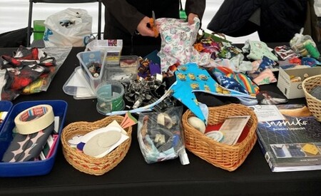 Ett bord fullt med pysselmaterial, korgar, tejp, tygbitar och småsaker. En person arbetar med händerna.