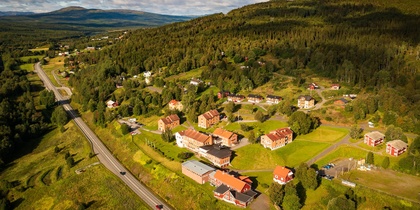 Flygfoto över en liten by med hus, vägar och grönskande landskap vid en skogsklädd kulle.