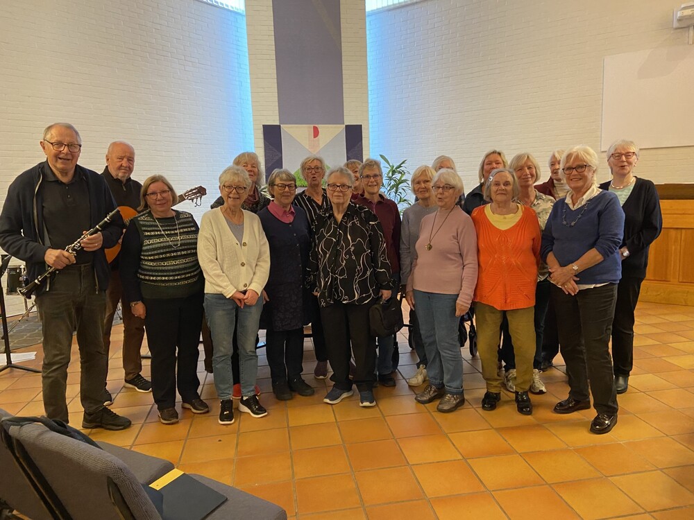 En grupp äldre personer står tillsammans inomhus, en man håller en gitarr. De ser glada ut.