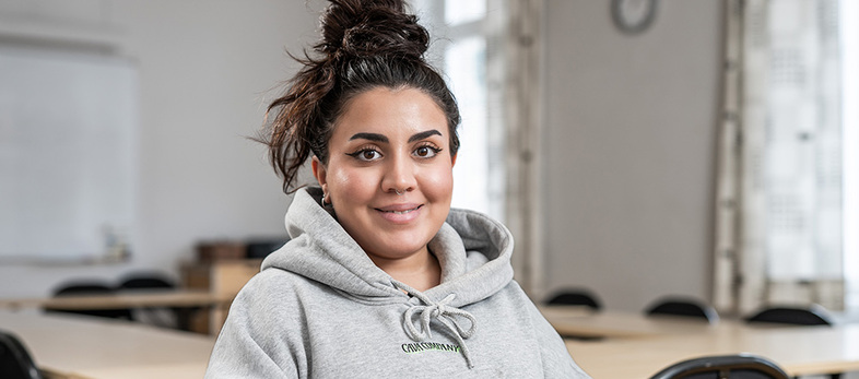 Ung kvinna med mörkt hår i knut och grå huvtröja sitter i ett klassrum och ler mot kameran.