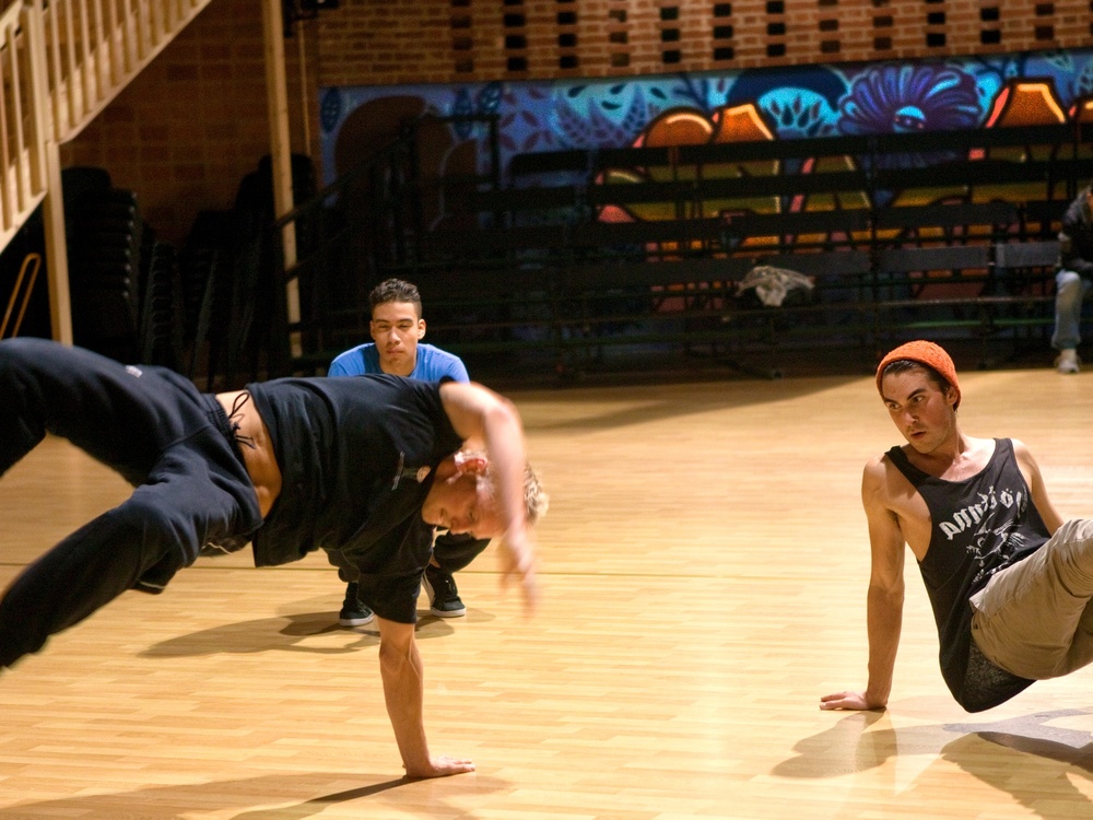 Tre unga personer dansar breakdance i en dansstudio med trägolv och graffitimålning på väggen.