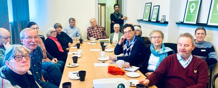 En grupp vuxna sitter runt ett konferensbord med kaffekoppar och anteckningar, i ett ljust mötesrum.