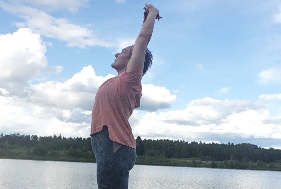 Person står vid en sjö och sträcker armarna mot himlen under en blå himmel med moln.