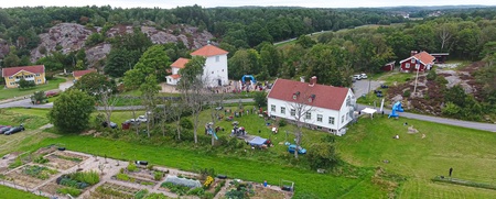 Lantlig gård med odlingslotter, vita hus och människor samlade utomhus, omgiven av grönska och skog.
