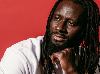 Pa Modou Badjie i vit t-shirt och svarta arbetsbyxor sitter på högtalare mot röd bakgrund