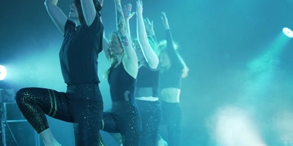 Fyra personer i glittriga byxor dansar på scen med armarna uppsträckta i blått scenljus.