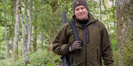 Man i jaktkläder står i en grön skog med ett gevär över axeln och tittar mot kameran.
