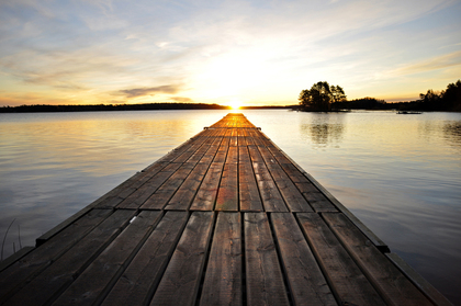 En lång brygga leder ut över en stilla sjö mot en solnedgång med träd i horisonten.