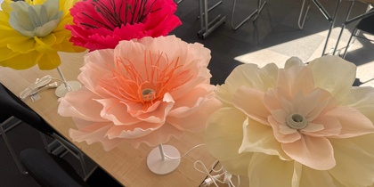 Fyra stora, färgglada konstgjorda blommor på lampfötter står på ett bord i ett mötesrum.