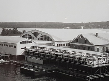Svartvit bild av en industribyggnad vid vattnet med stor skorsten och bryggor.