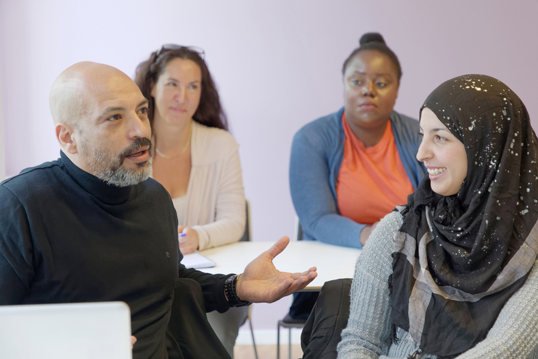 Fyra personer sitter runt ett bord och samtalar i ett ljust rum. En kvinna bär hijab och ler.