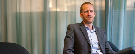 Man i kostym sitter avslappnat i en fåtölj framför ett blågrönt draperi.