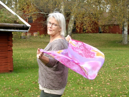 Äldre kvinna med glasögon står utomhus och håller en rosa sjal som blåser i vinden.