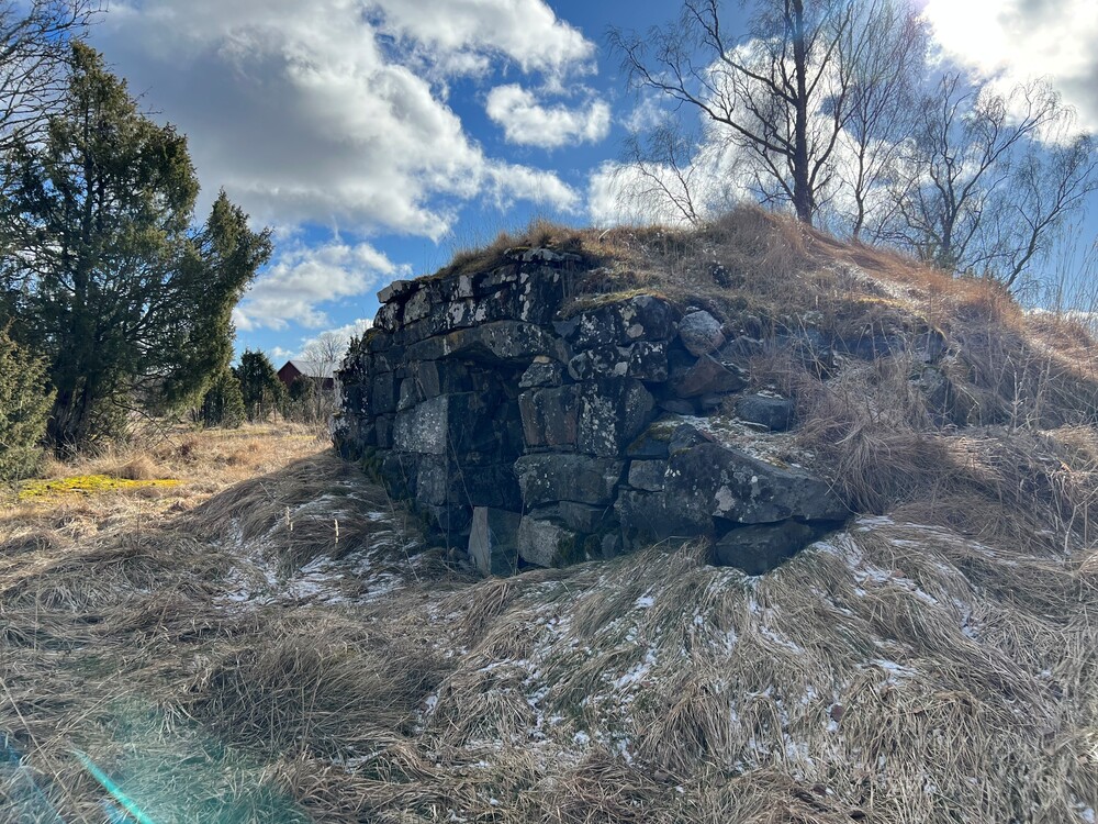 En gammal stenkällare täckt av gräs och mossa i ett öppet landskap under en blå himmel med moln.